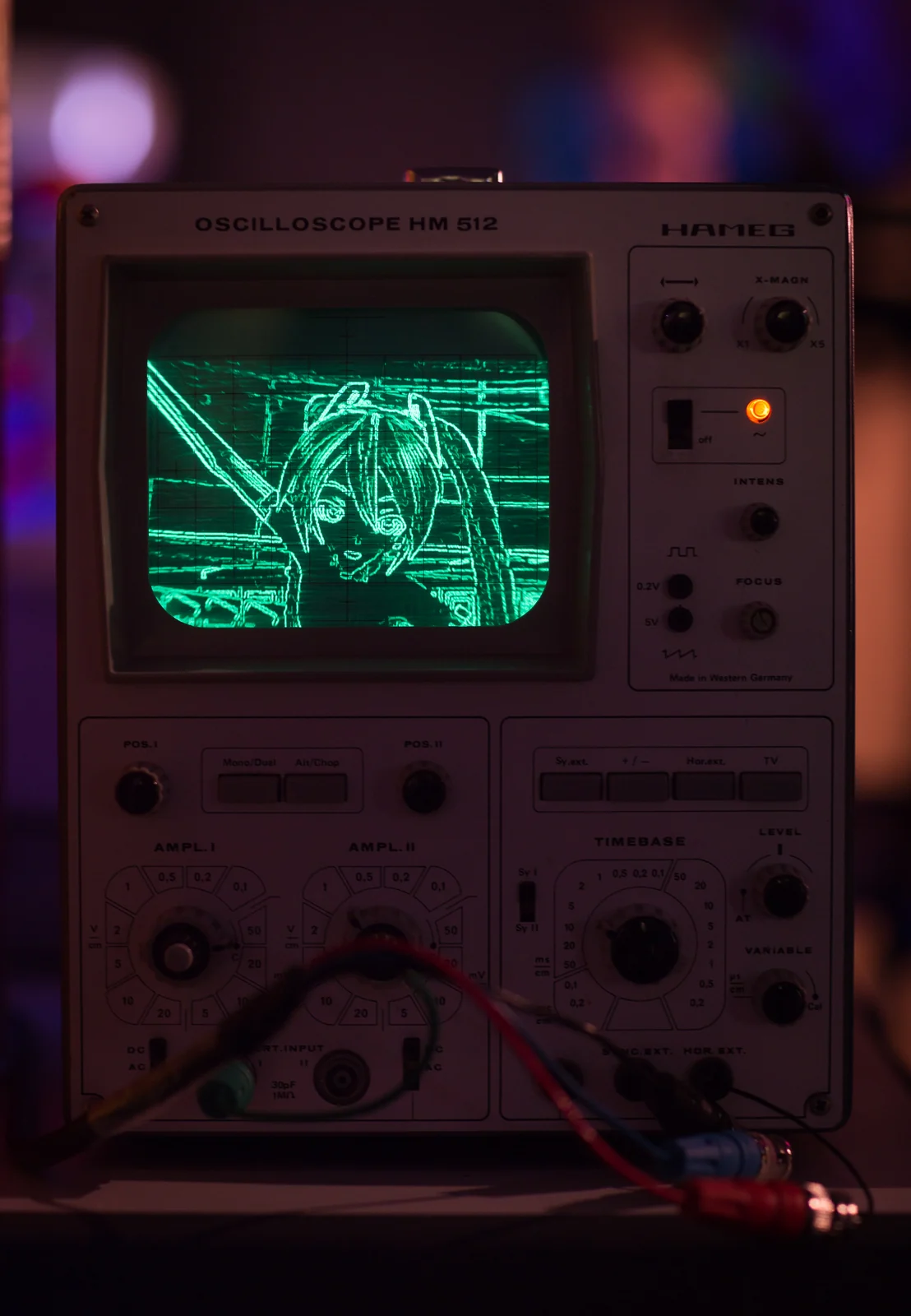 An analog oscilloscope displaying a drawn figure, with controls and cables below.